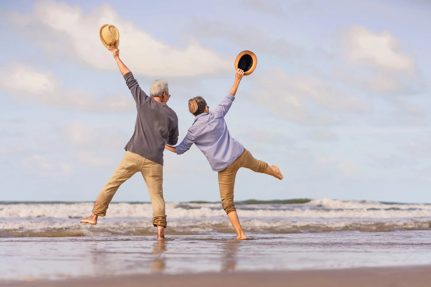 Two people, an elderly man with gray hair and a woman with brown hair in a bun, are dancing and jumping on the beach near the ocean, holding hats in the air.