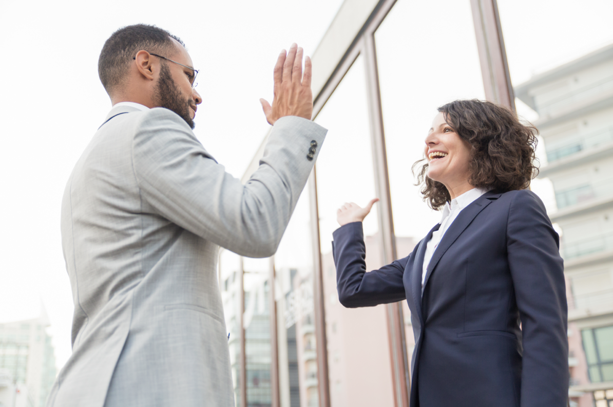 A man and a woman in business attire giving each other a high five outside on a city street.