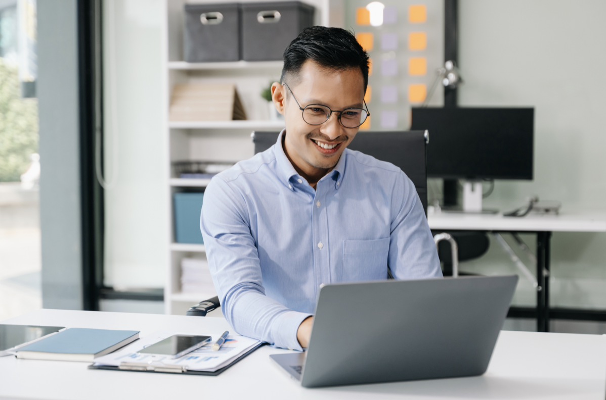 A man sitting at a desk using a laptop, smiling, in an office with shelves and a window in the background.