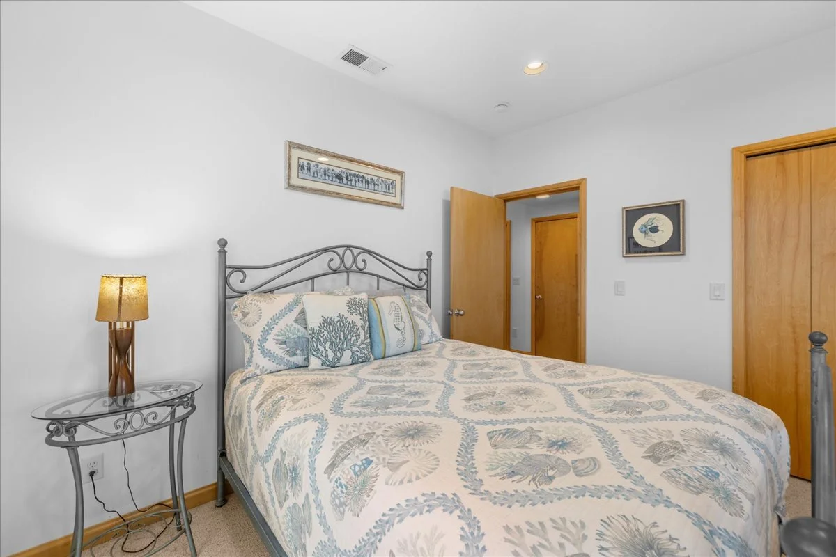 A bedroom with a bed, pillow, and colorful bedspread, a side table with a lamp, two closed wooden doors, a framed picture of a blue fish on the wall, and a framed photo above the bed.