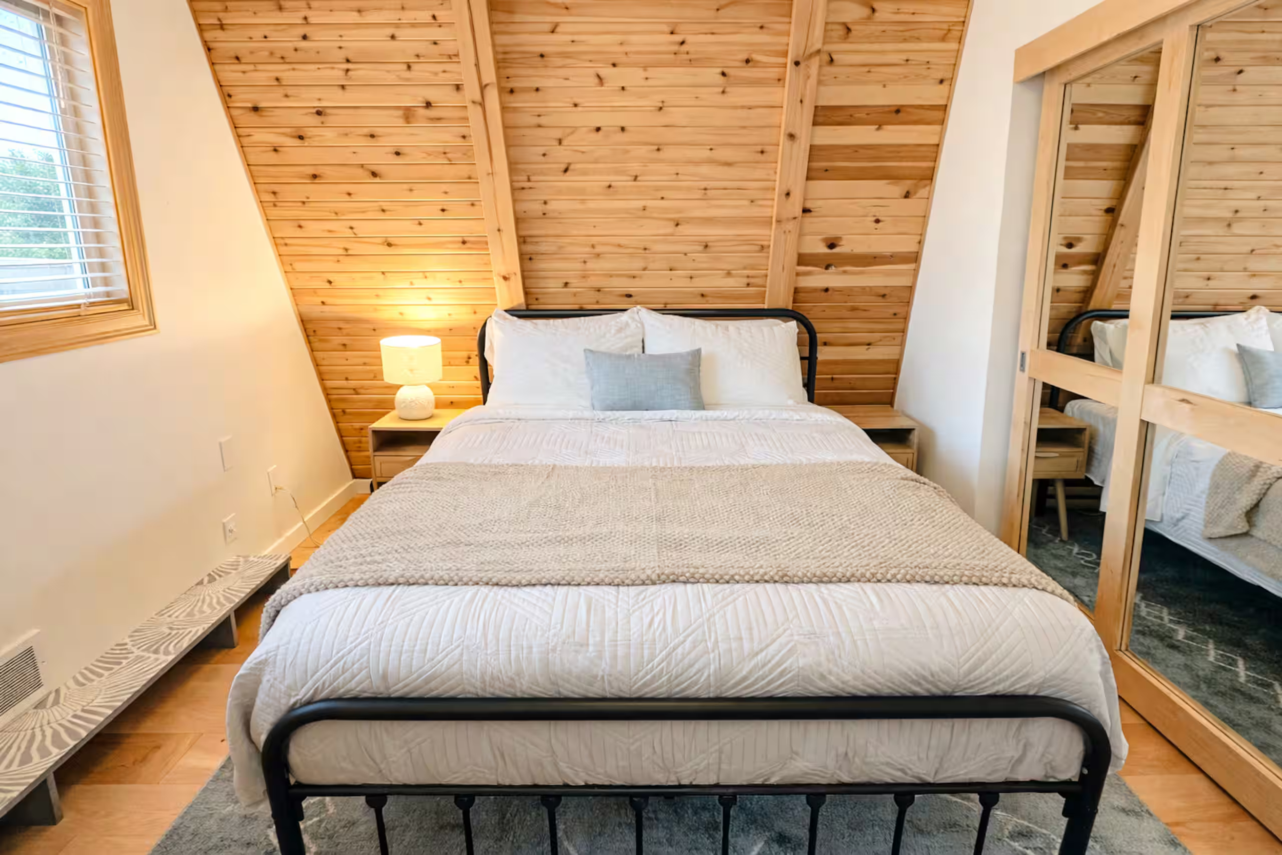 A bedroom with a wooden slanted ceiling, two beds with white bedding and pillows, a nightstand with a lamp, a window with blinds, and a mirrored closet door.