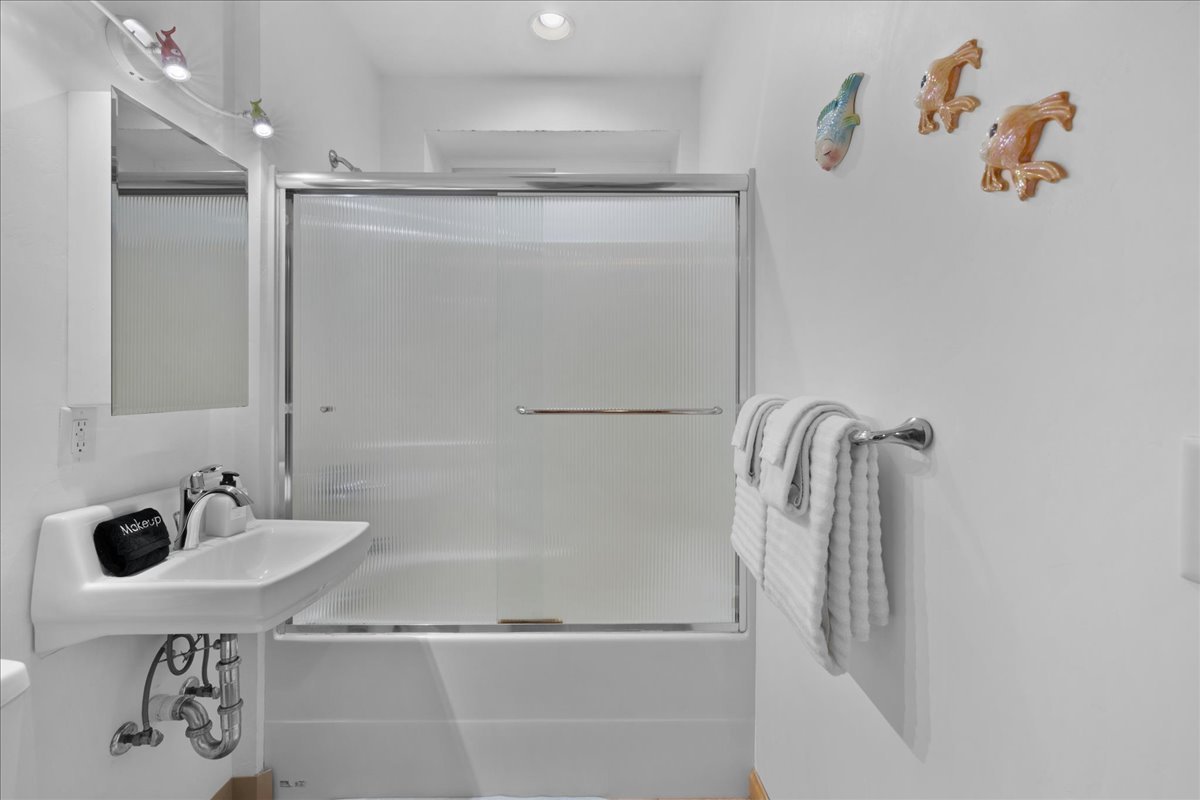 Small white bathroom with a sink, mirror, and frosted glass shower door, decorated with fish and sea-themed wall art, and white towels hanging on a towel rack.