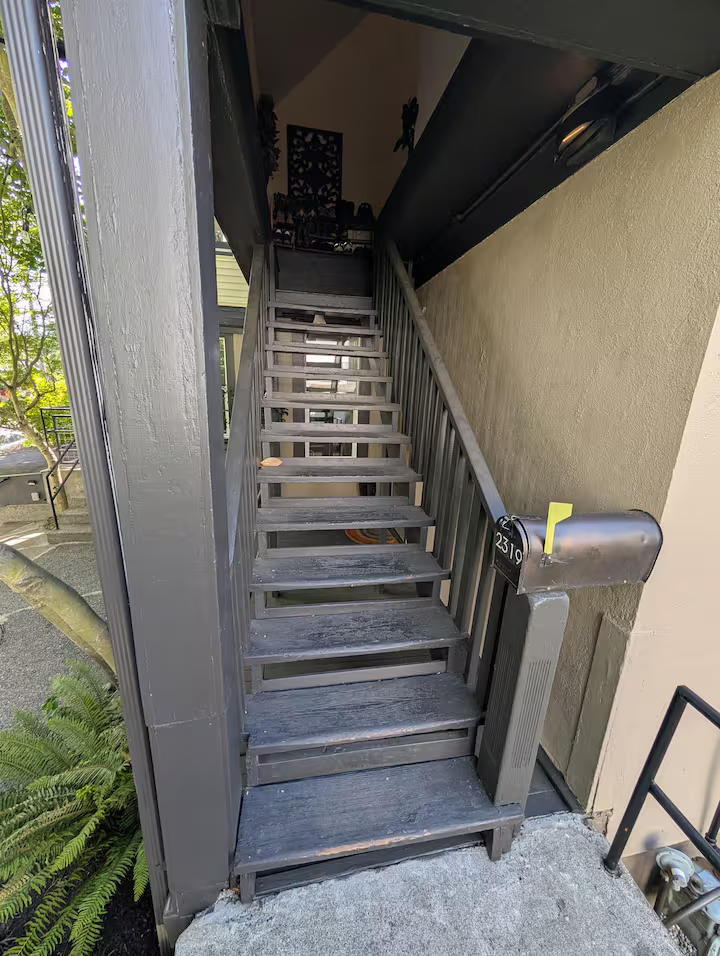 Outdoor wooden staircase leading up to a second-floor entrance, with a black mailbox labeled '2519' mounted on the right side and a small black railing on the right. Some greenery and plants are visible to the left.
