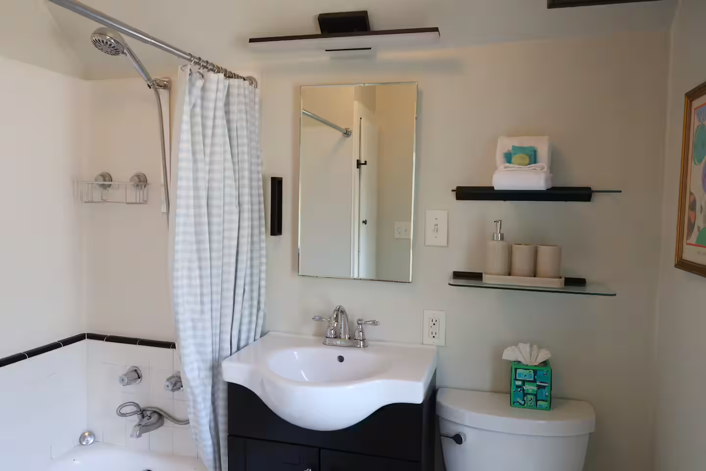 A small bathroom with a shower on the left side with a curtain, a white sink with a black cabinet, a mirror above the sink, a toilet with a tissue box on top, and two glass shelves holding towels and cleaning supplies.