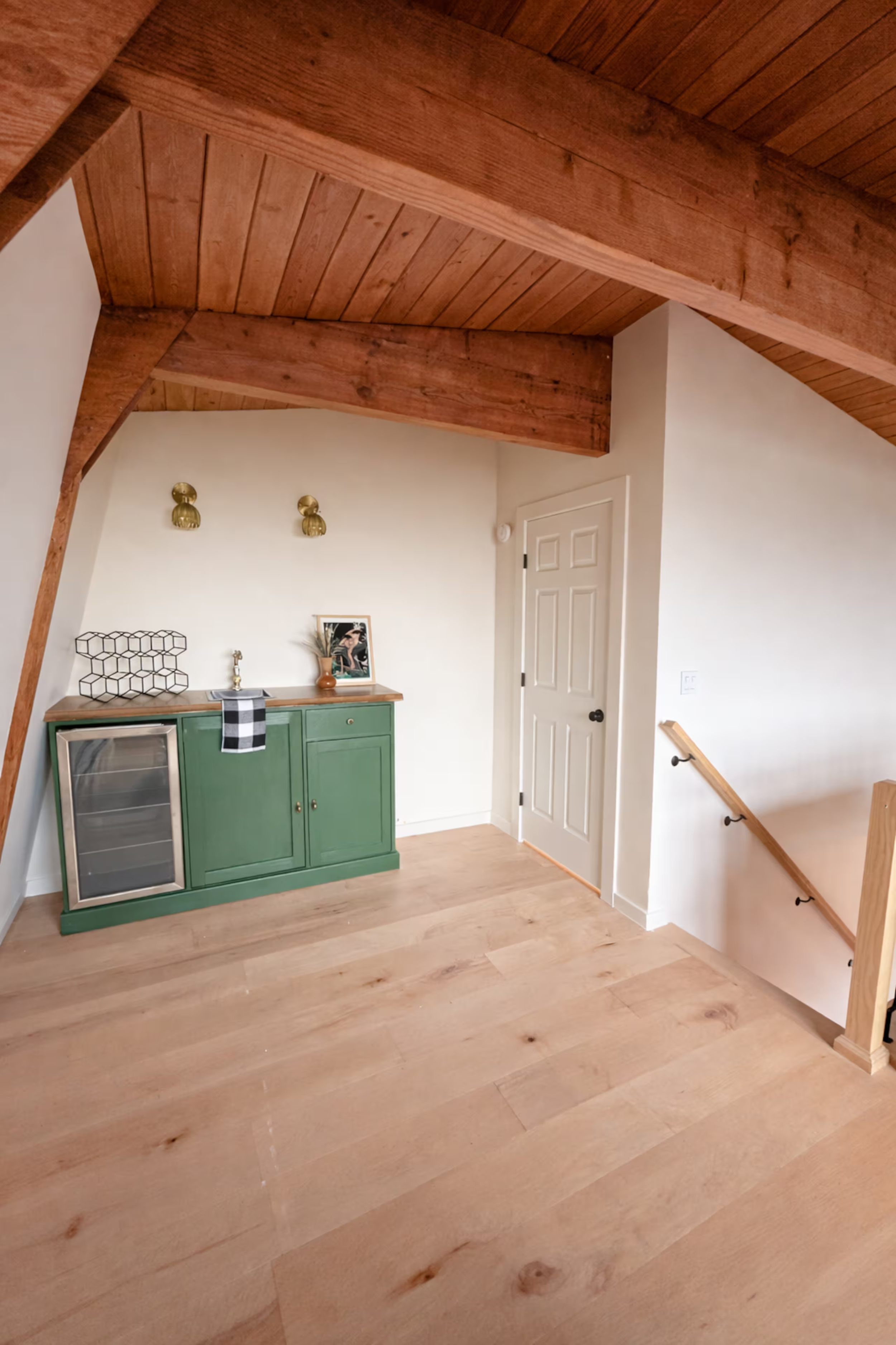 Interior view of a room with wooden ceiling and floors, green cabinet, framed photo, decorative objects, and staircase with dark railing.