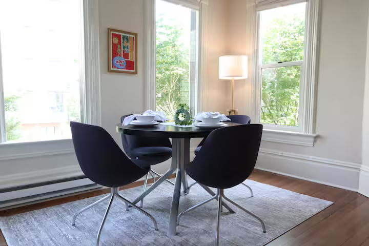 A round black dining table set for four with plates and napkins, surrounded by four black chairs, situated on a light-colored rug in a room with large windows, a floor lamp, and framed artwork on the wall.