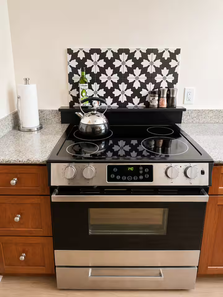 Kitchen stove with four burners, a kettle on the left front burner, and a black backsplash with white floral pattern. The stove has a digital display showing 4:40. To the left is a roll of paper towels, and to the right are salt and pepper shakers an