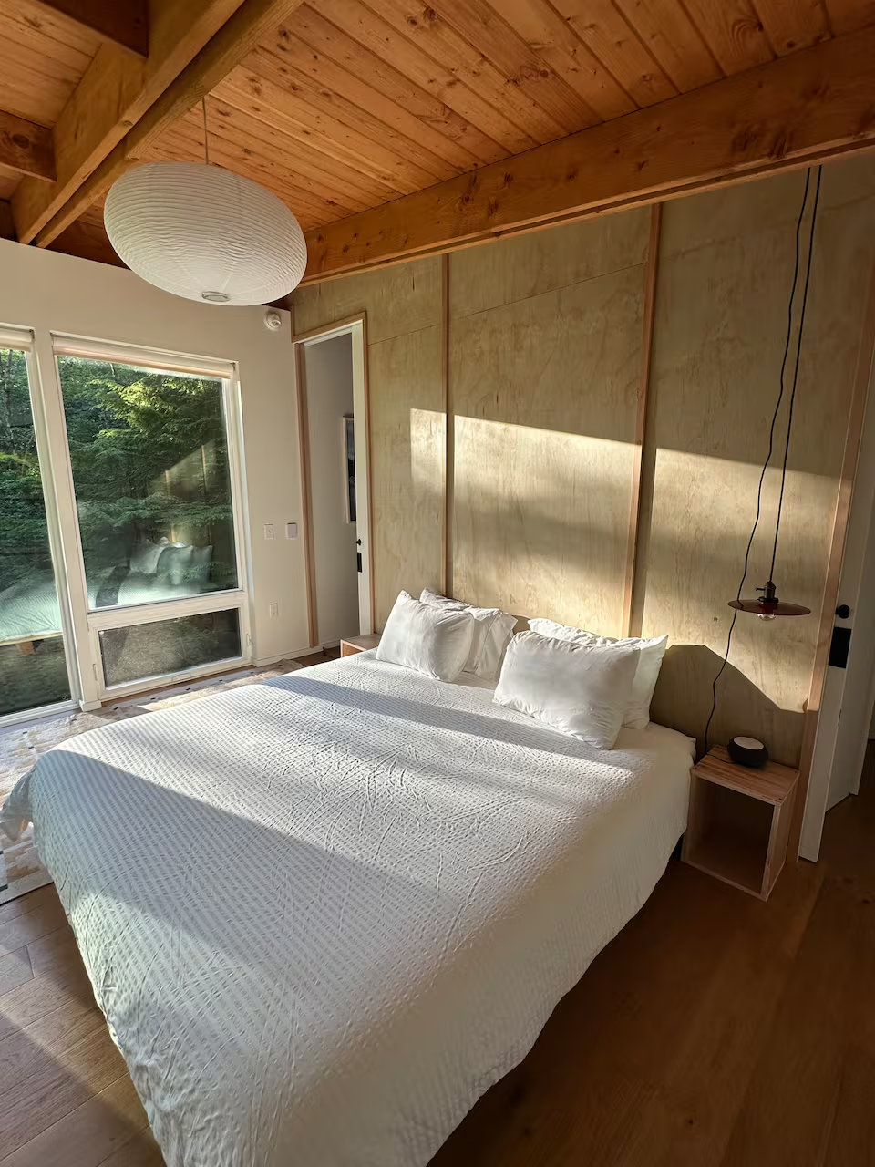 A bedroom with a wooden ceiling, sliding glass door, and a bed with white bedding and pillows. Sunlight is streaming onto the bed, and there is a small nightstand with a black speaker and a hanging pendant light beside the bed.