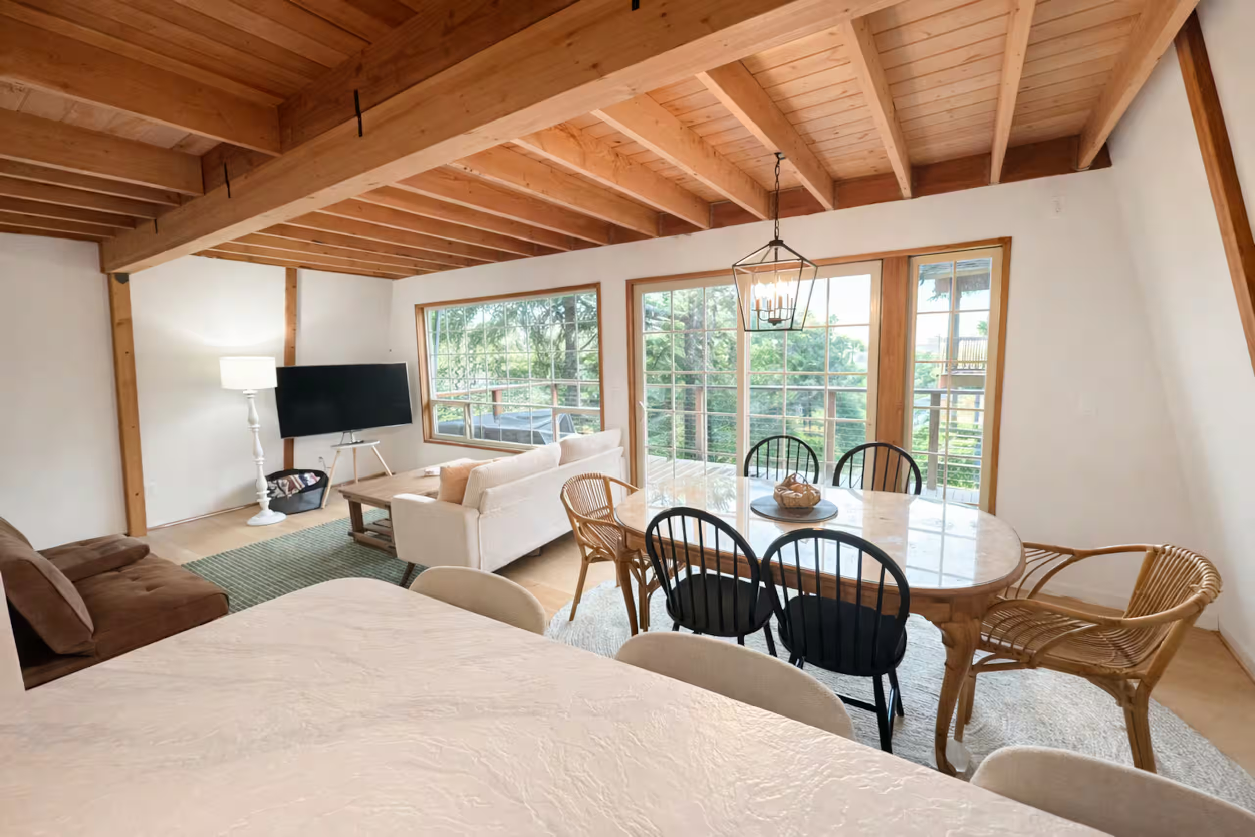 Living room with wooden vaulted ceiling, large windows, a flat-screen TV, a white sofa, a wooden dining table with wicker and black chairs, and a chandelier.