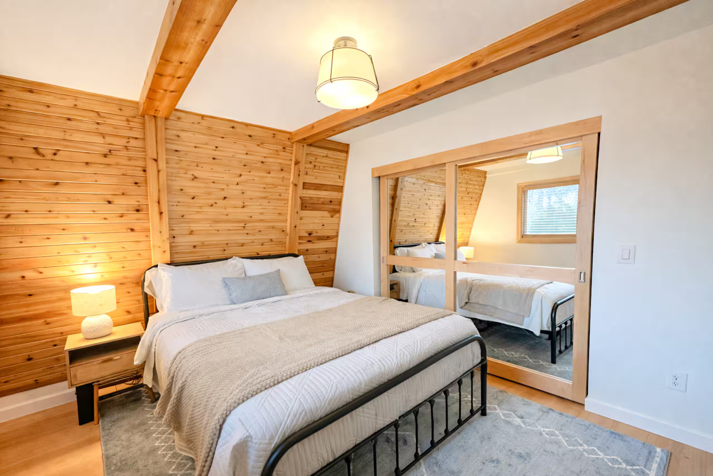 A bedroom with a rustic design featuring a bed with white bedding and a decorative pillow, a nightstand with a textured white lamp, and a mirror reflecting part of the room. The room has wooden beams and paneling on the walls and ceiling, a window wi