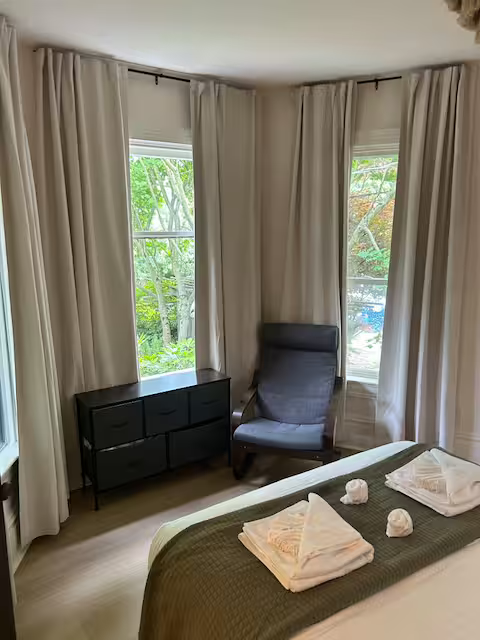 A bedroom with three large windows, cream curtains, a black dresser, a dark gray armchair, and a bed with white towels and toiletries on top.