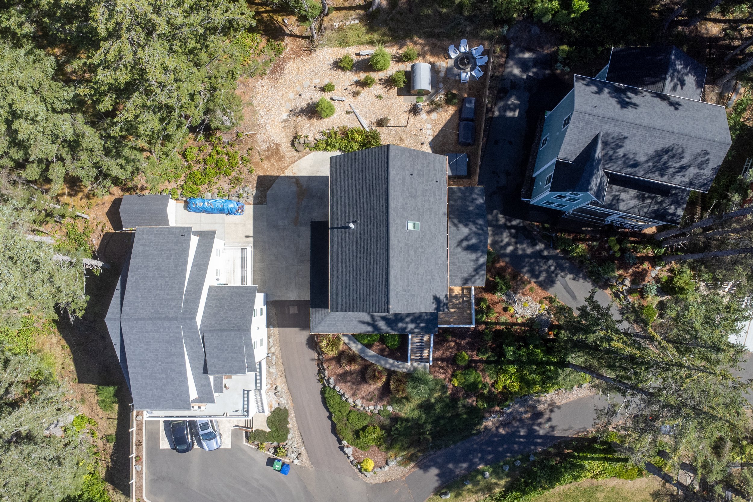 An aerial view of a residential property surrounded by trees, showing several houses, parked cars, a backyard with outdoor furniture, and a curved driveway.