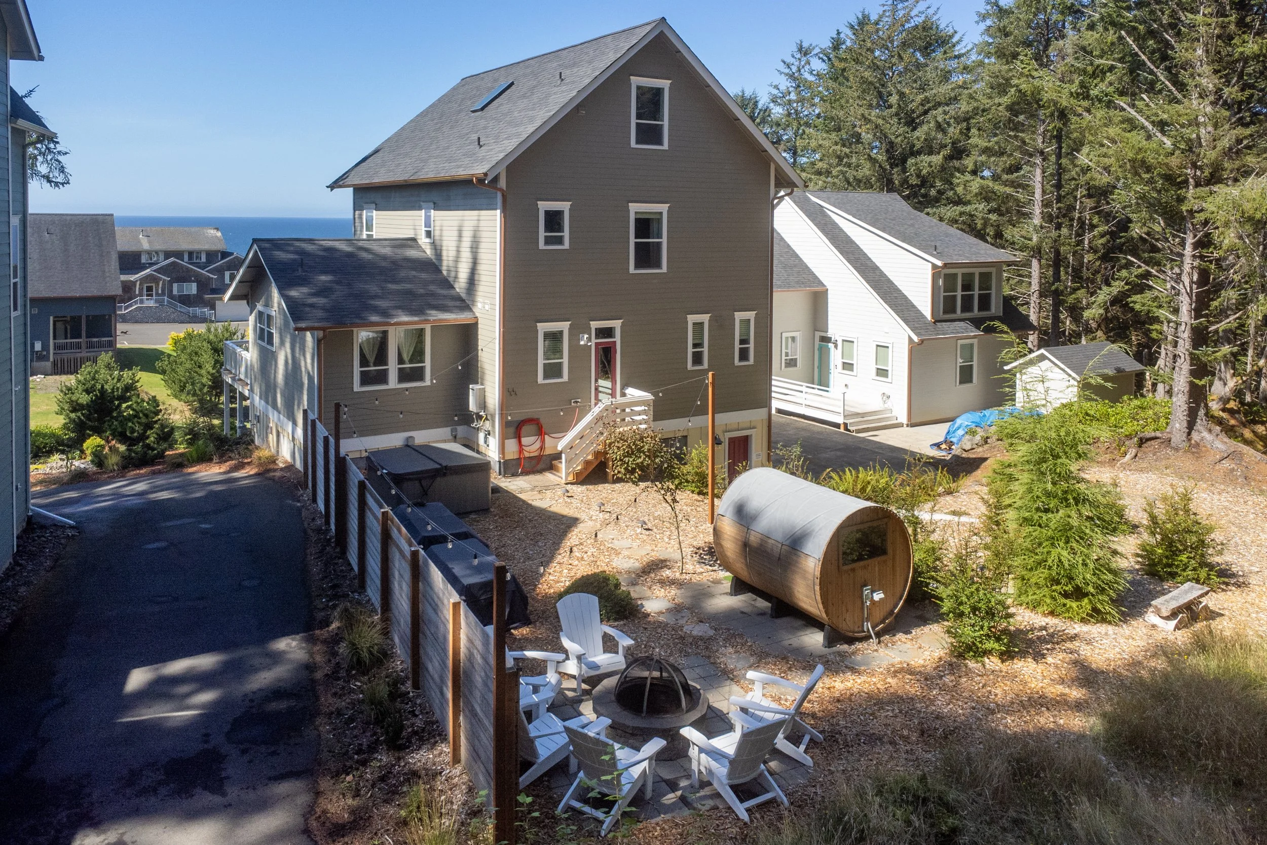 View of a multi-story house with beige and white siding, a gray roof, and multiple windows, situated on a hillside with trees and a view of the ocean in the background. There is a backyard with a fire pit surrounded by white chairs, a small shed, and