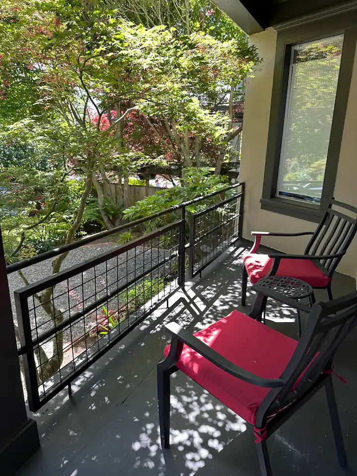 Balcony with two black chairs with pink cushions, outside view of trees and greenery, sunlight casting shadows, window on the right.