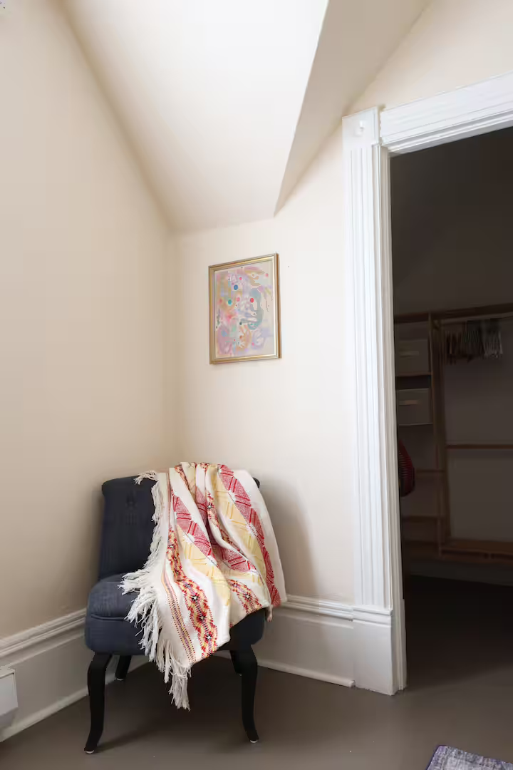 A cozy corner with a dark blue upholstered armchair draped with a cream blanket with red and yellow patterns, positioned against cream-colored walls and a white ceiling with crown molding. A framed abstract artwork hangs above the chair, and part of 