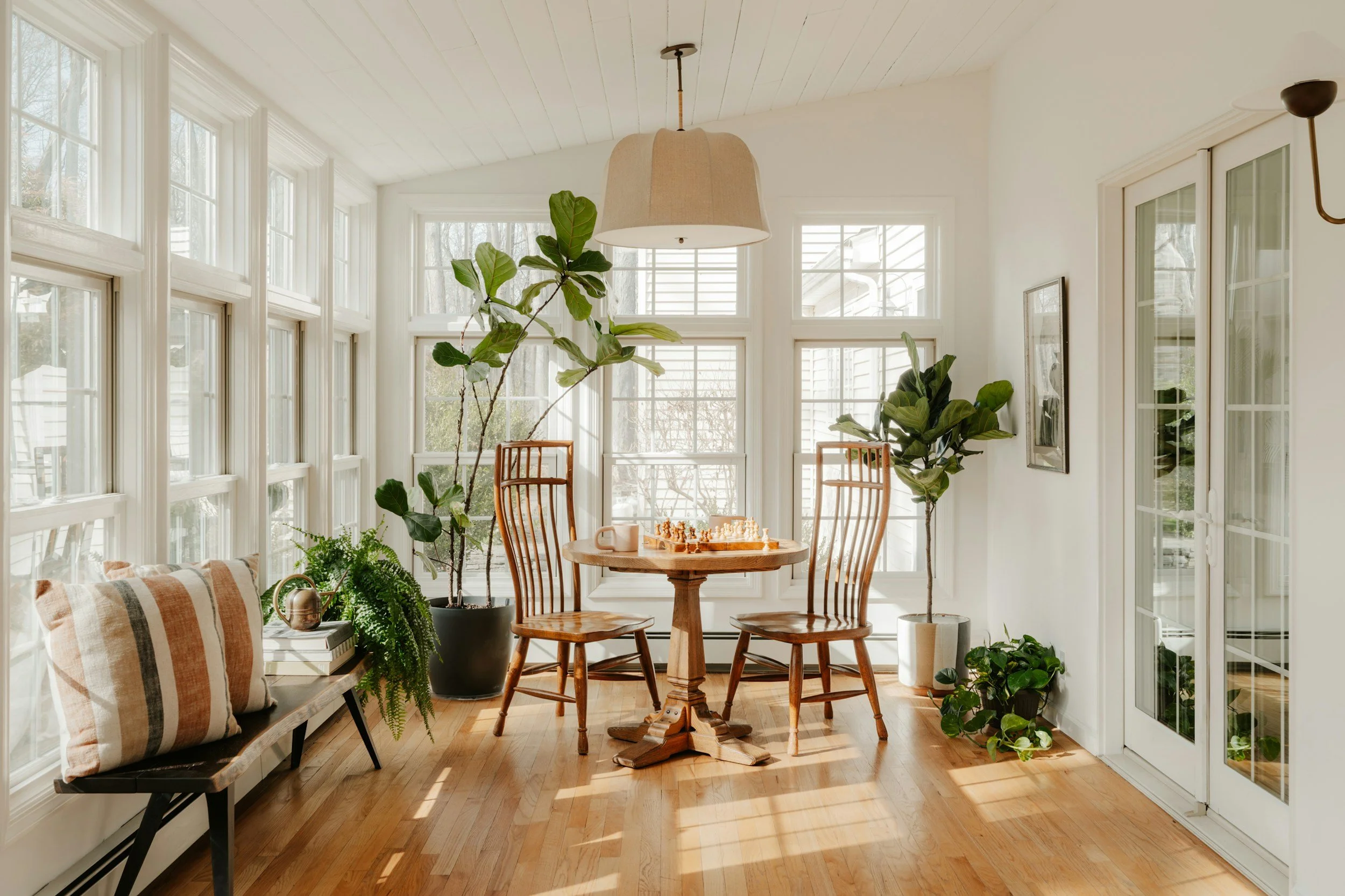 Bright sunroom with large windows, wooden floor, round wooden table, wooden chairs, potted plants, and a bench with plaid cushions.