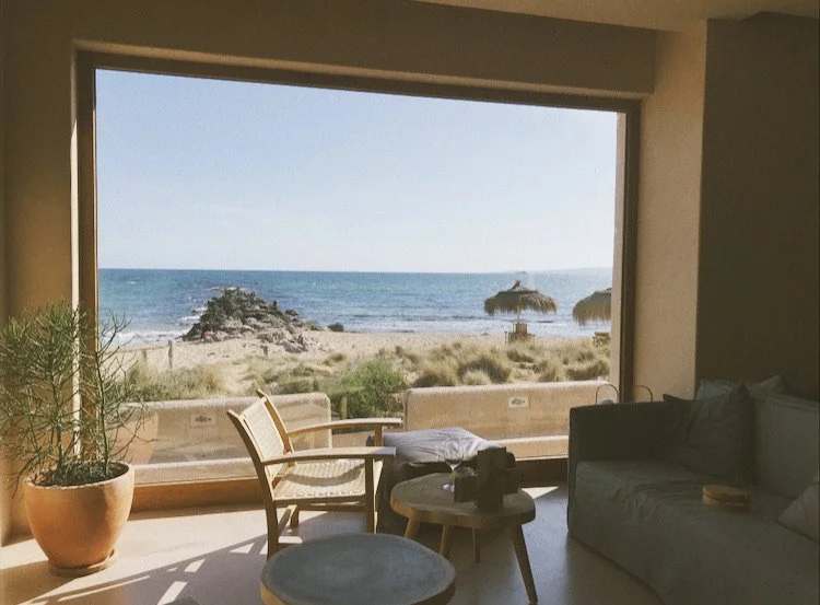 Beach view from a living room with a large window, showing sand, grass, rocks, and ocean, with beach chairs and umbrellas outside.