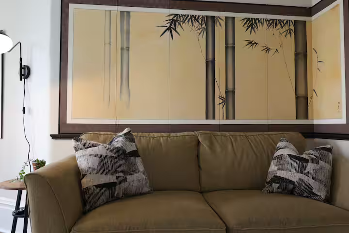 Living room with beige sofa, three decorative pillows, a bamboo-themed wall art, and a small side table with a plant in a pot.