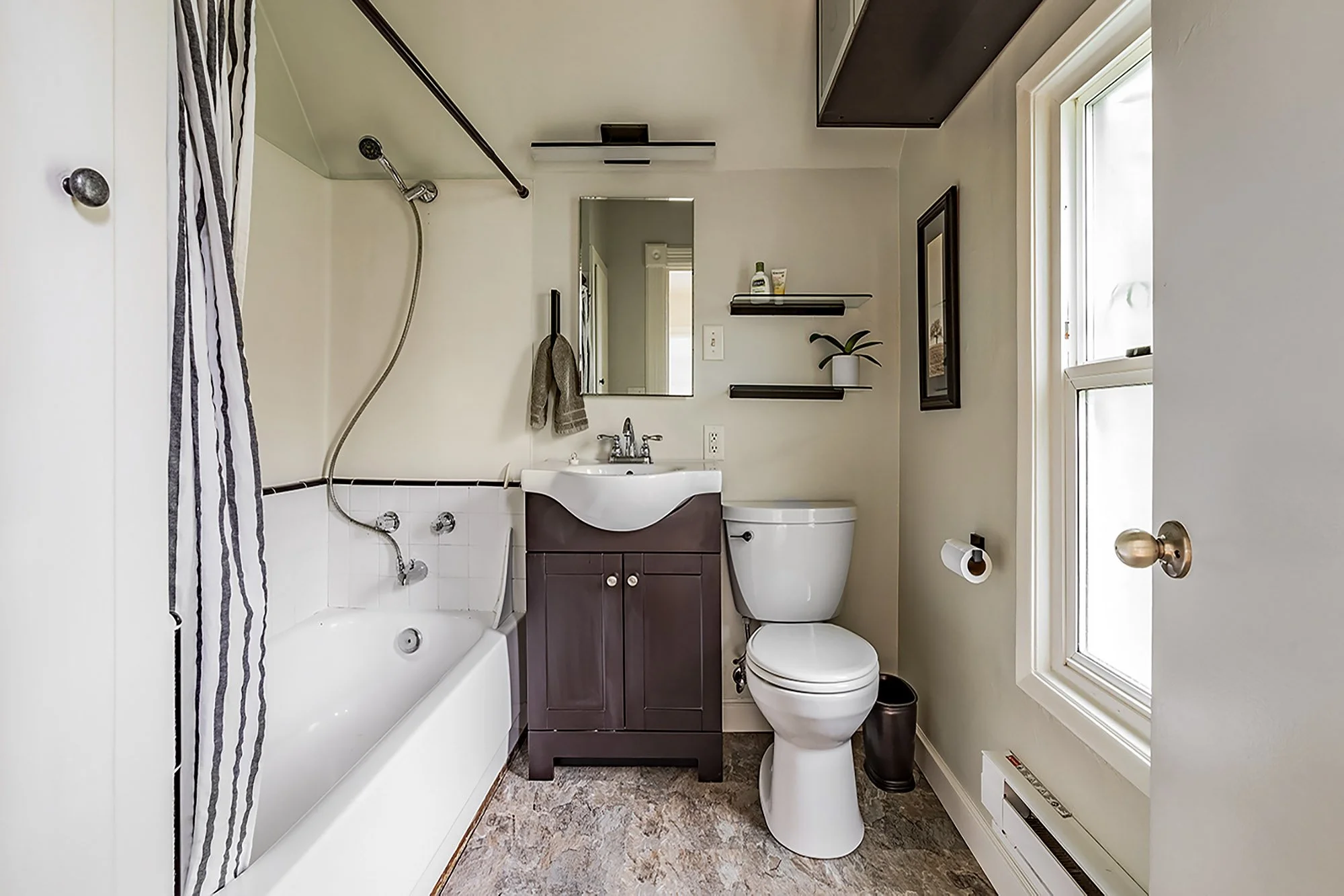 A compact bathroom with a bathtub and shower, a mirror above the sink, a toilet, and decorative shelves with a plant and toiletries, bright natural light from large windows.