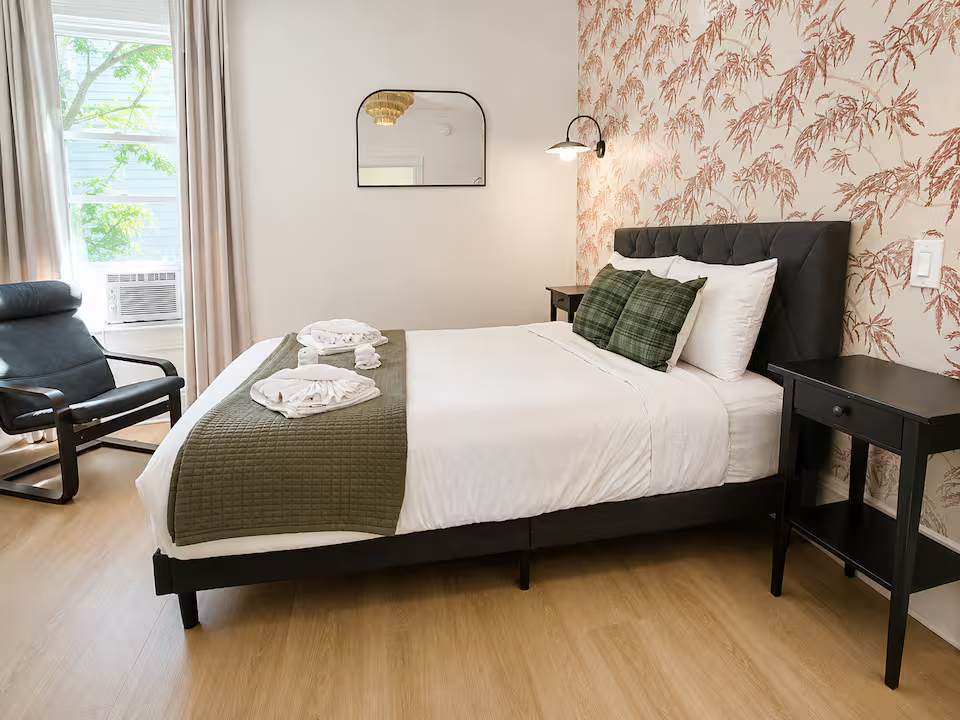 A cozy bedroom with a bed featuring white sheets, green plaid pillows, and a green quilted blanket. There are folded towels on the bed. Next to the bed is a black nightstand. A large window with beige curtains and a window air conditioning unit allow