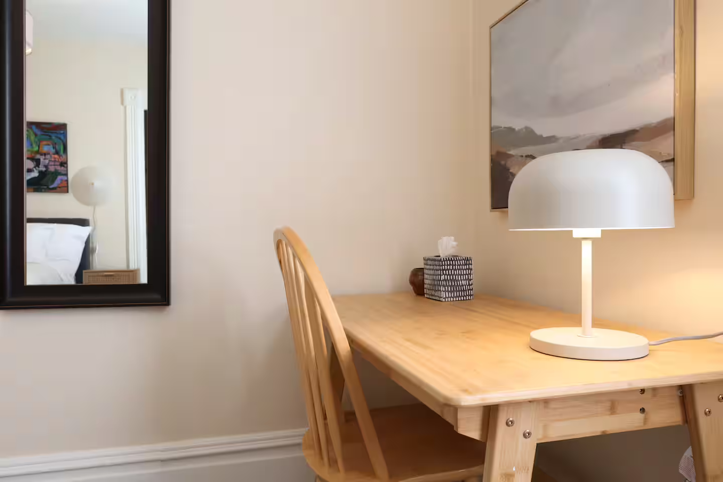 A wooden desk with a white table lamp, tissue box, and small decorative items in a cozy room. A large landscape painting hangs on the wall above the desk. A mirror reflects part of an adjacent room with a bed and colorful artwork.