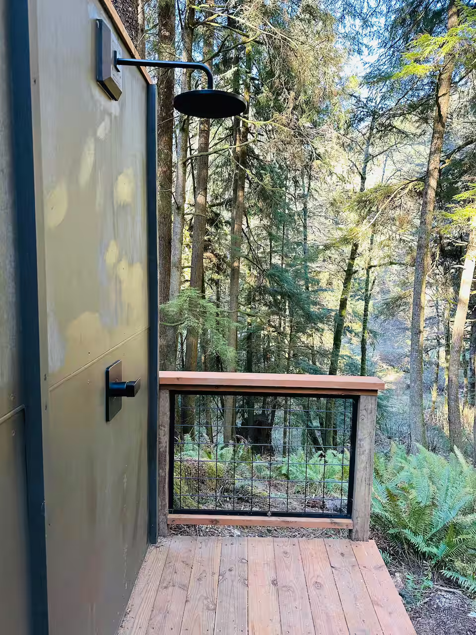 A wooden outdoor shower with a showerhead mounted on a dark green wall, overlooking a forest with tall trees and green foliage.