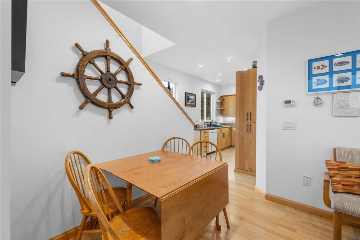 Dining area with a wooden table and four wooden chairs, nautical decor including a ship's wheel on the wall, kitchen in the background with wooden cabinets, a window, and wall-mounted art with fish images.