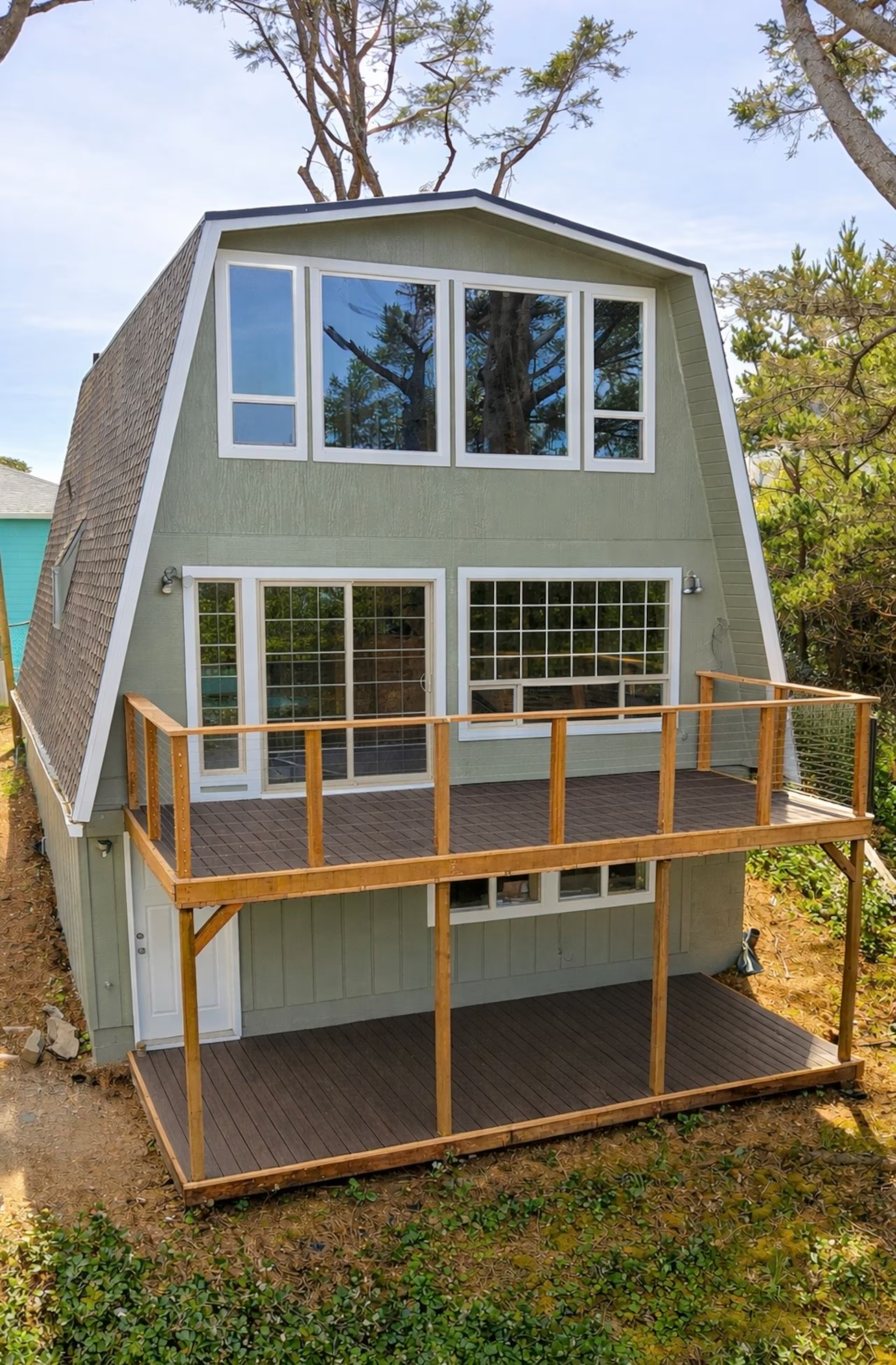 A three-story house with green exterior, large front windows, and a wooden deck on the second floor. The house has a unique angled roof and is surrounded by trees and greenery.