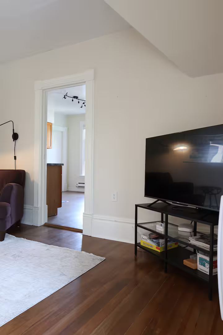 Living room with a large flat-screen TV on a black stand, a purple chair, a white area rug, and view into the kitchen through a doorway. The room has hardwood floors and white walls.