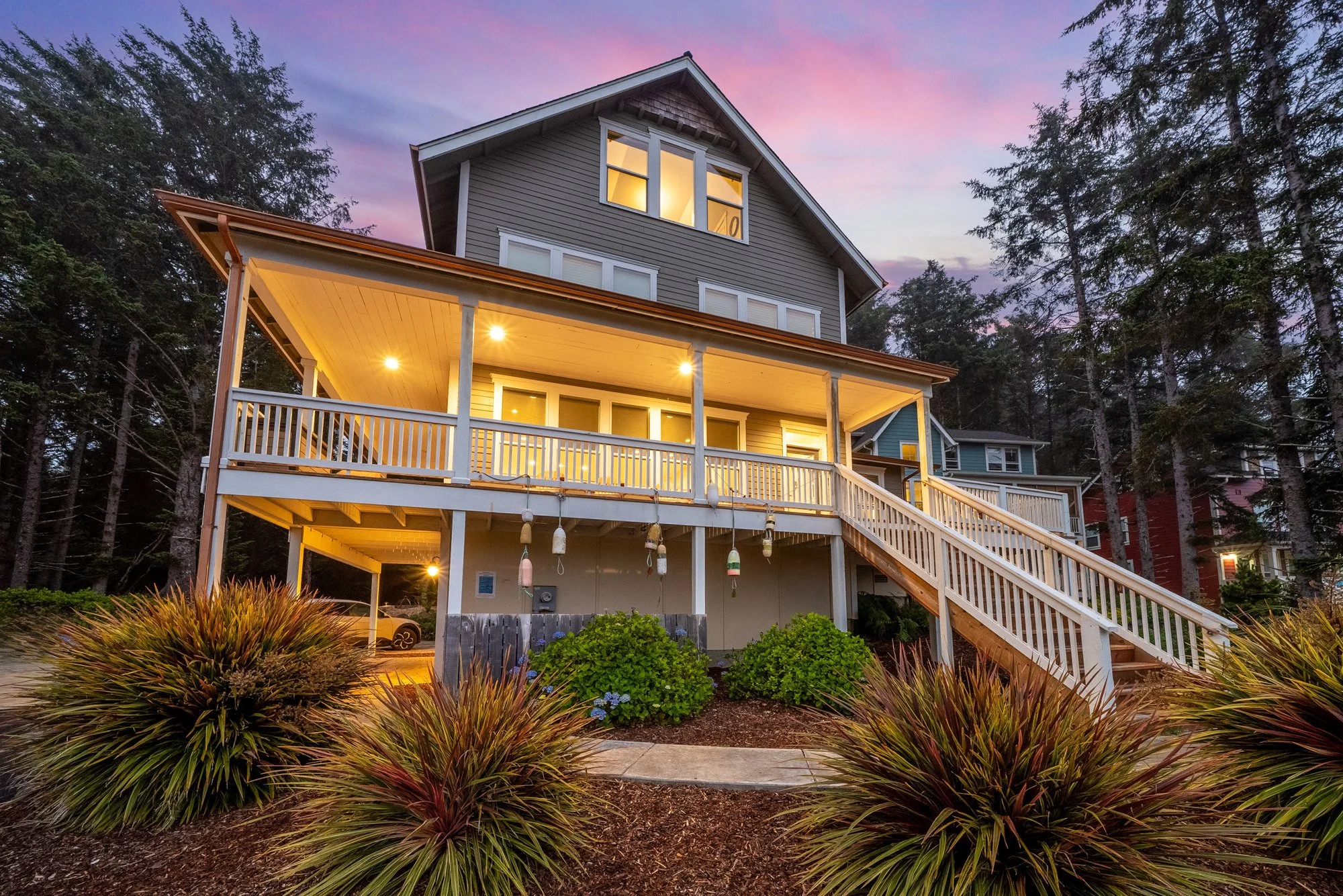 A multi-story house with a spacious porch, surrounded by trees. The house has gray siding, white trim, and a brown roof. The porch lights are on, illuminating the area, and the sky is a mix of pink and purple during dusk. There are landscaped bushes and plants in the foreground.