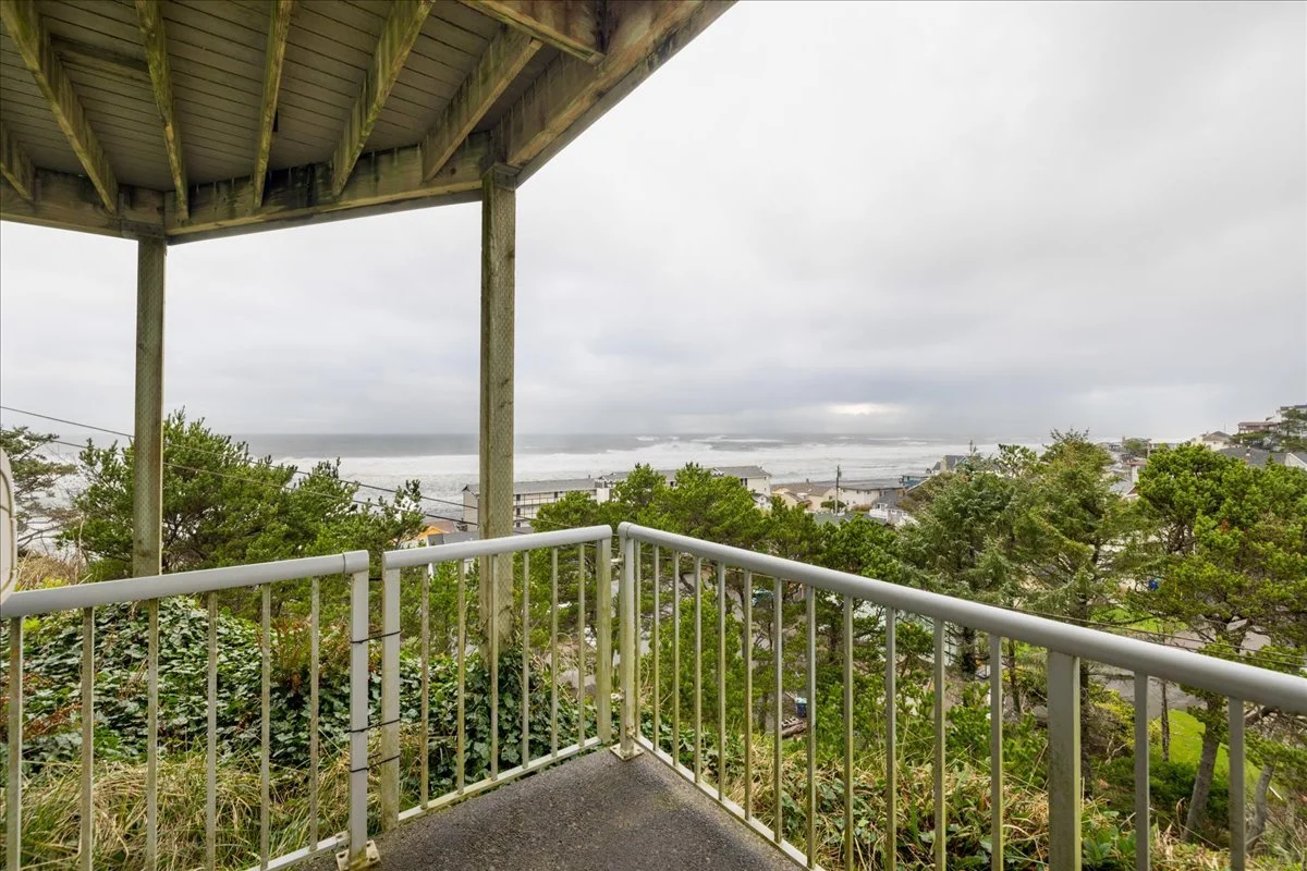 View from a balcony overlooking a coastal town with trees, houses, and the ocean in the distance on a cloudy day.