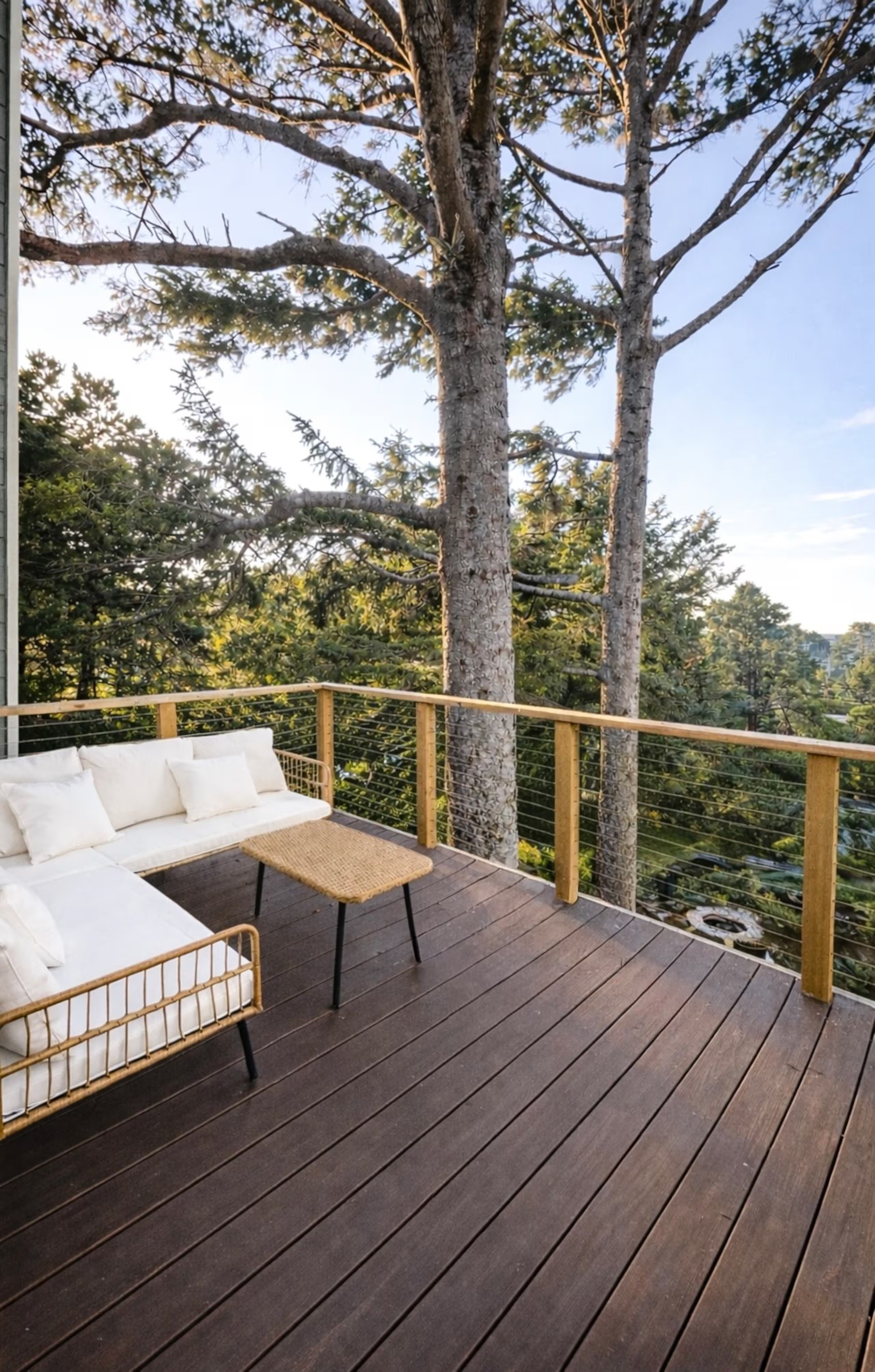 A wooden outdoor balcony with white cushioned seating and a small wicker table, surrounded by a wooden railing, overlooking large trees and a cityscape in the distance during daytime.