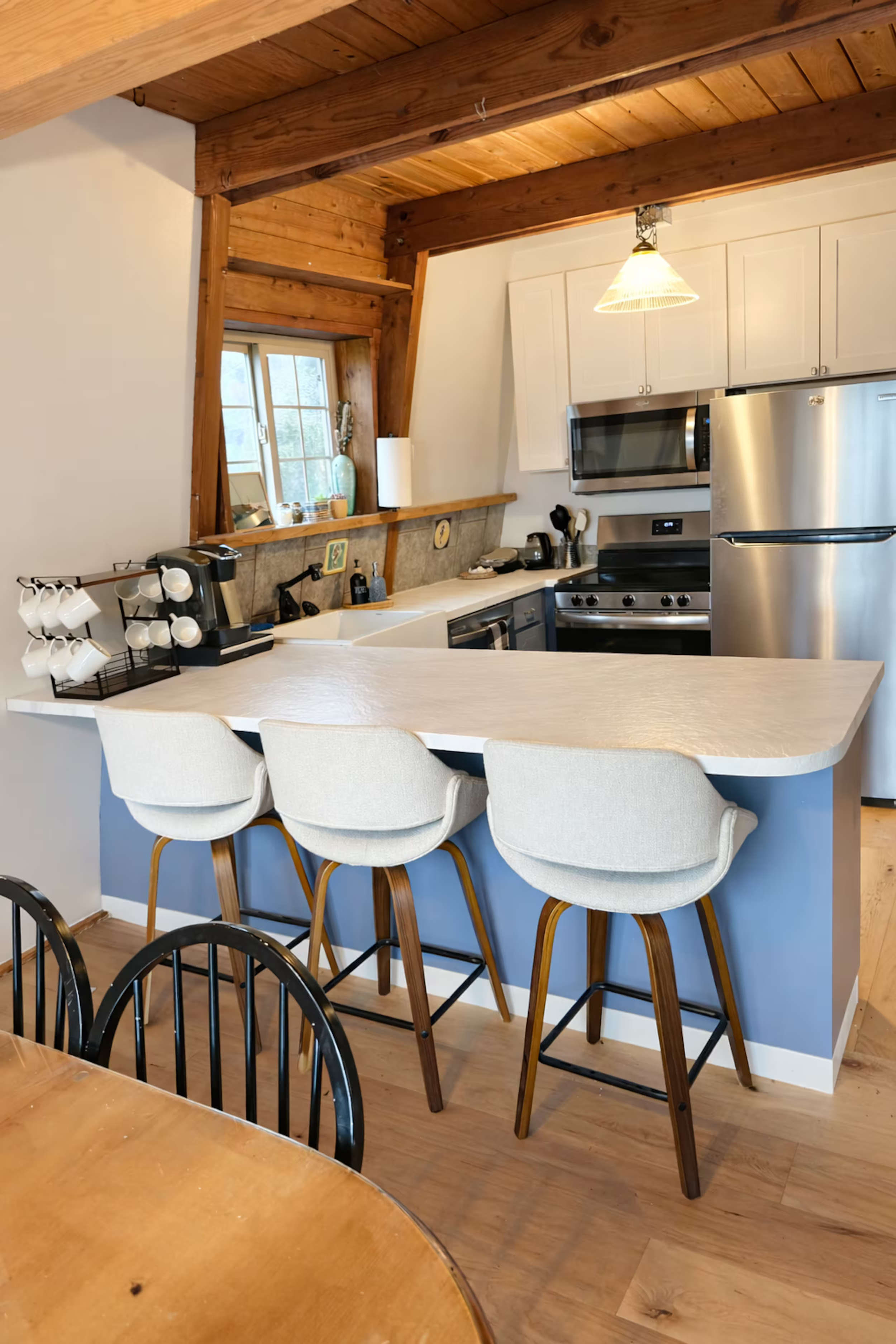 Kitchen with wooden beams on ceiling, white cabinets, stainless steel refrigerator, microwave, black stove, countertops with kitchen essentials, and a window with a small countertop and decorative items.