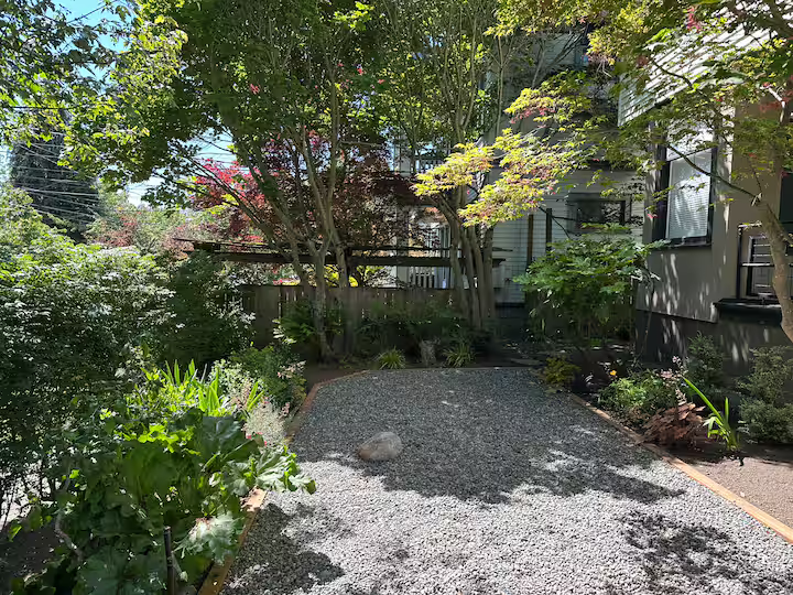 A gravel pathway in a lush backyard garden with various trees, shrubs, and plants, and a house with windows in the background.