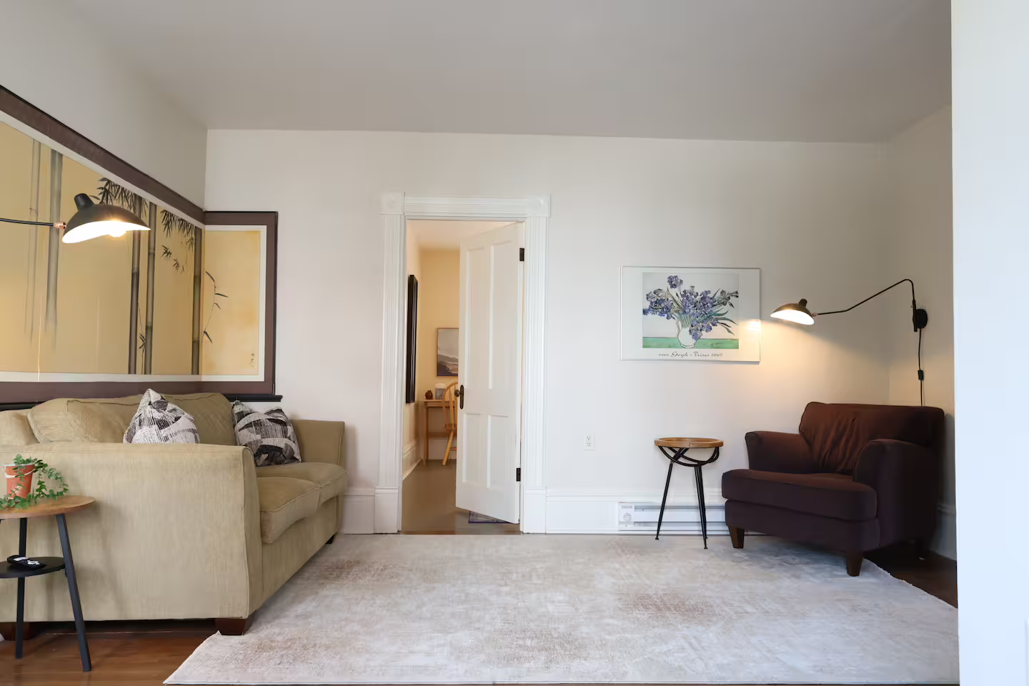 Living room with a beige couch, a dark red armchair, a small round side table, a wall-mounted light, framed artwork of purple flowers, and an open doorway leading to another room.