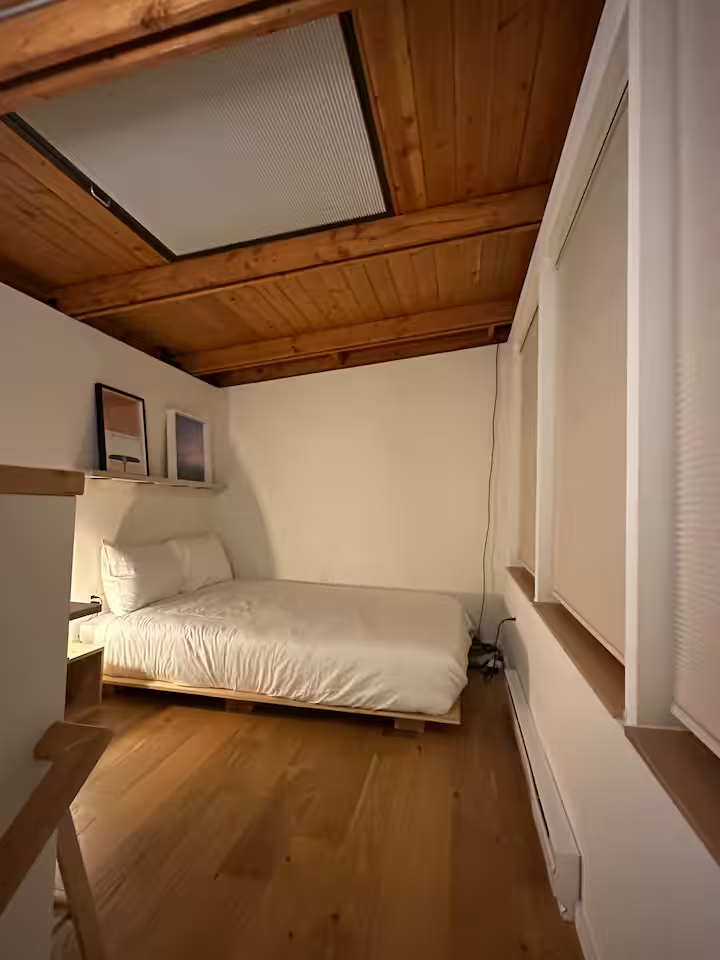 A cozy bedroom with a sloped wooden ceiling, a white bed with white bedding, a small wooden nightstand with a lamp, artwork on a shelf, and a window with blinds.