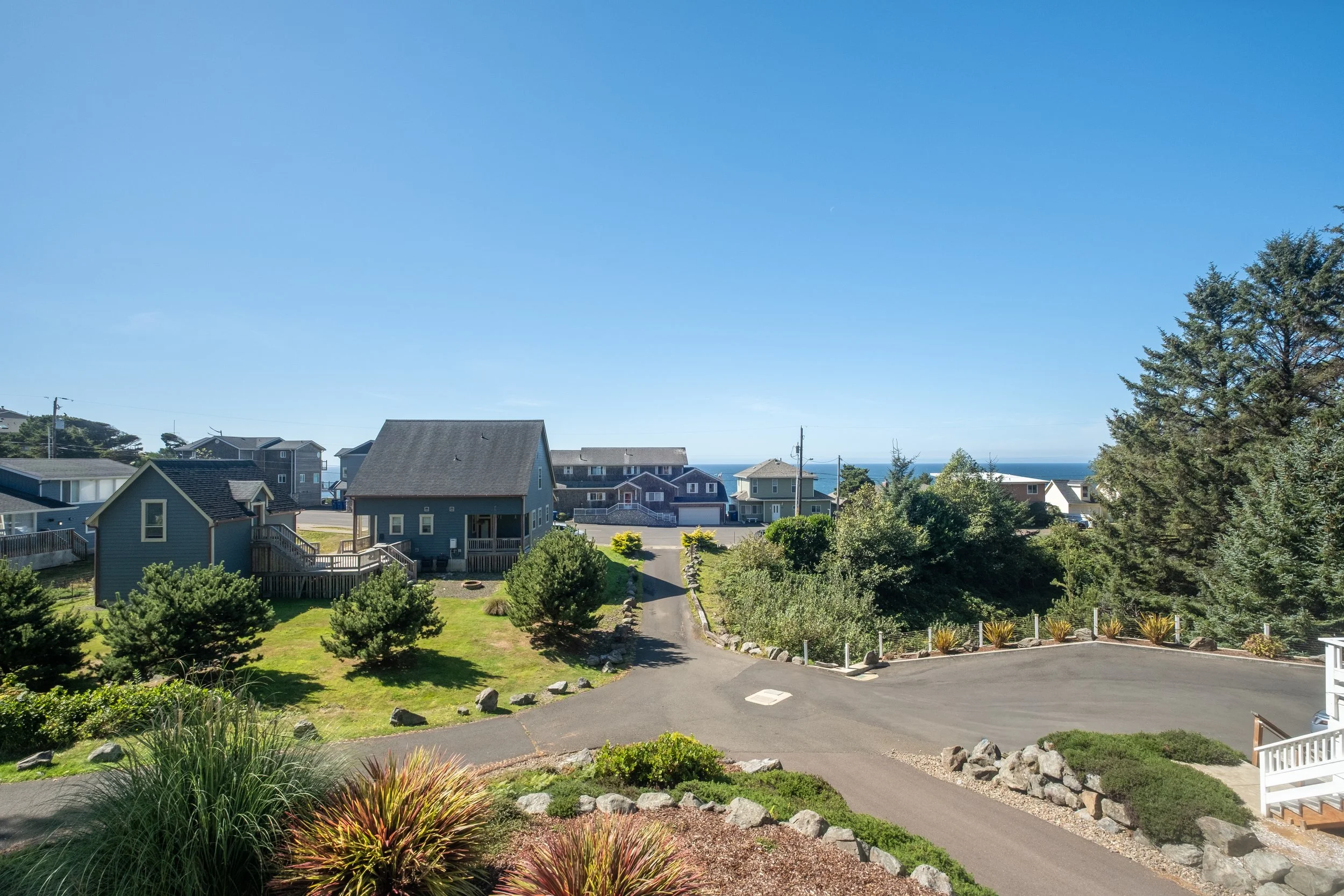A seaside residential neighborhood with several houses, lush greenery, and a clear view of the ocean under a bright blue sky.