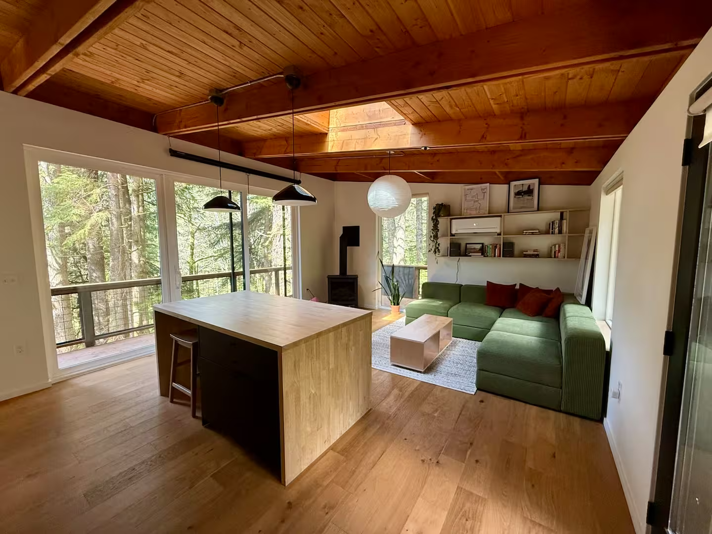 Living room with large windows showing a forest view, wooden ceiling, green sectional sofa with red pillows, white shelving with books, wooden coffee table, potted plant, and a wood stove.