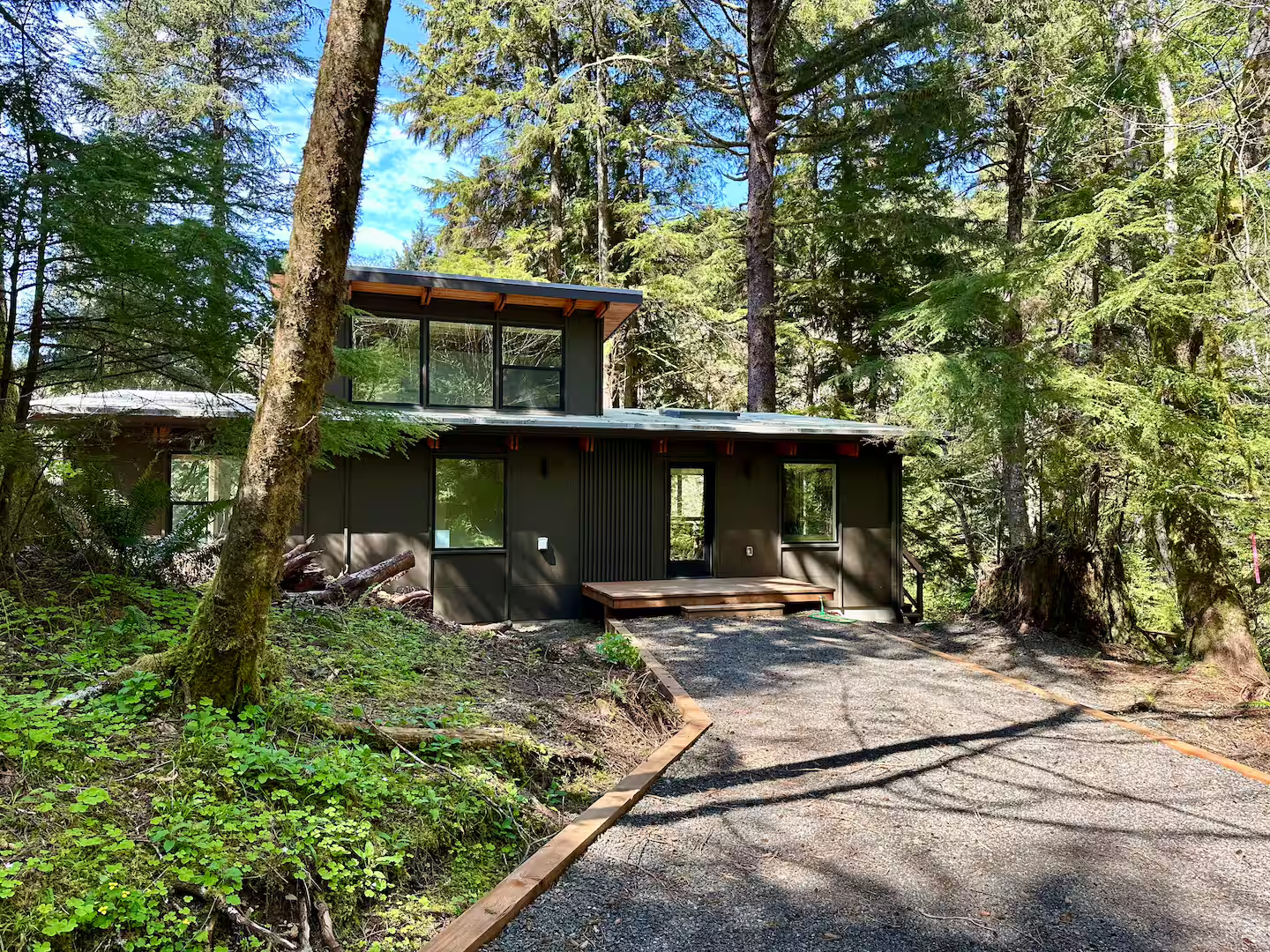 A modern house in a wooded area with tall trees, featuring dark exterior walls and large windows. The house has a small wooden deck at the entrance and a gravel driveway.