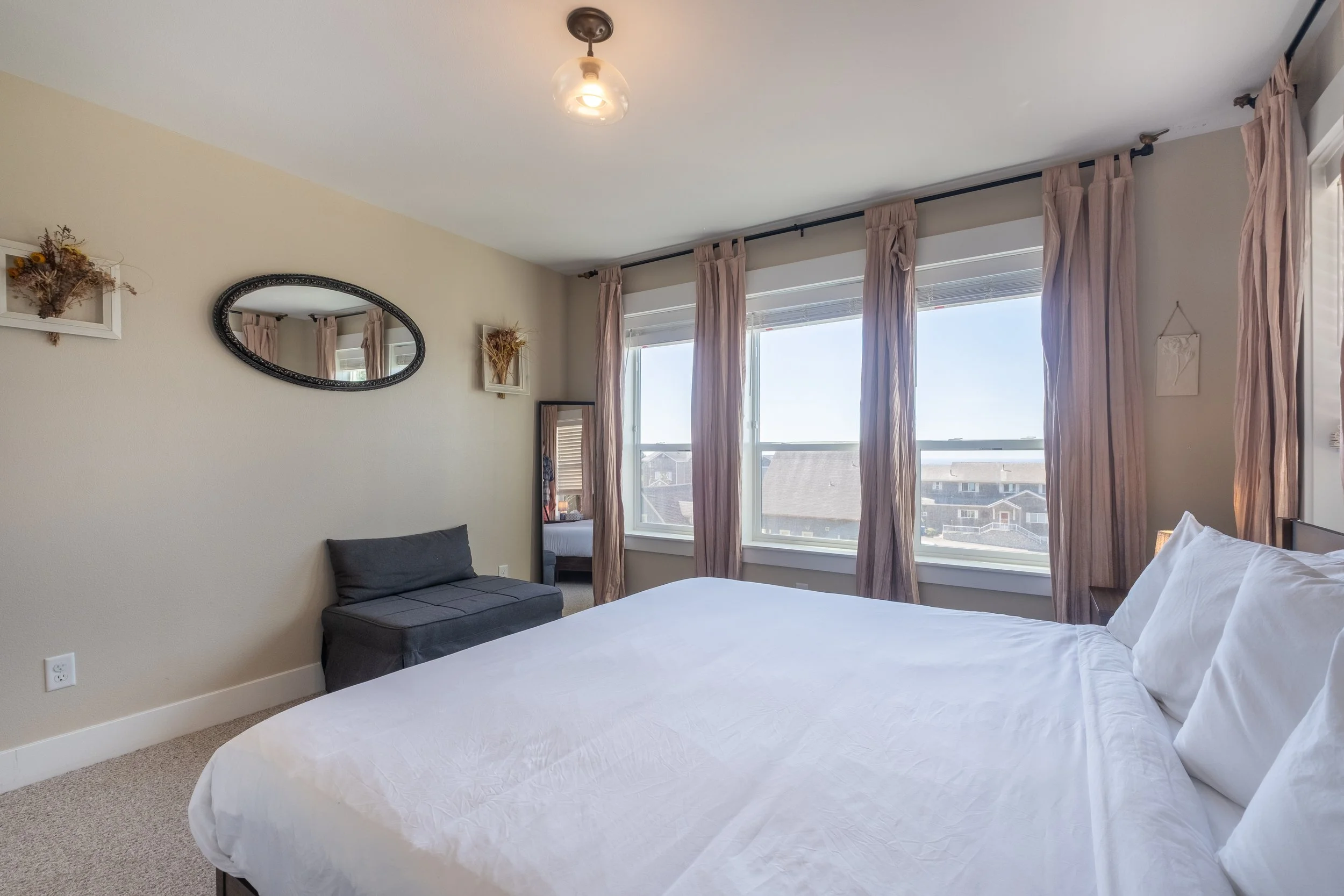 A bedroom with large windows covered by pink curtains, a white bed, a gray bench, a mirror, and wall decorations.