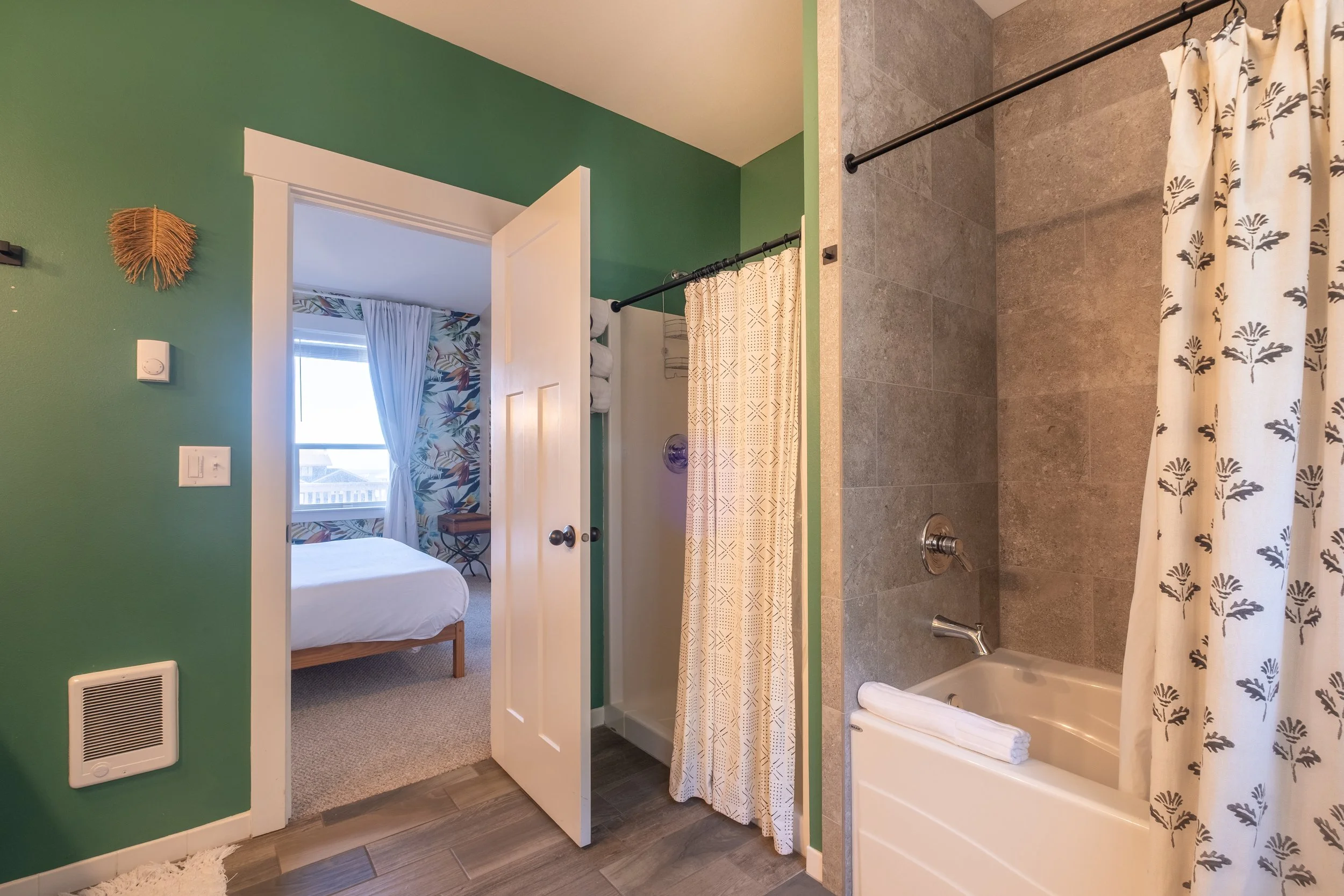 Bathroom with a bathtub, shower curtain with a leaf pattern, green wall, and view into a bedroom with a bed, window with curtains, and tropical wallpaper.
