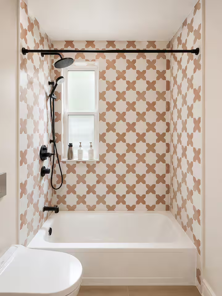 Bathroom with pink and white geometric patterned tile wall, white bathtub, black shower fixtures, a window, and three soap dispensers on the windowsill.