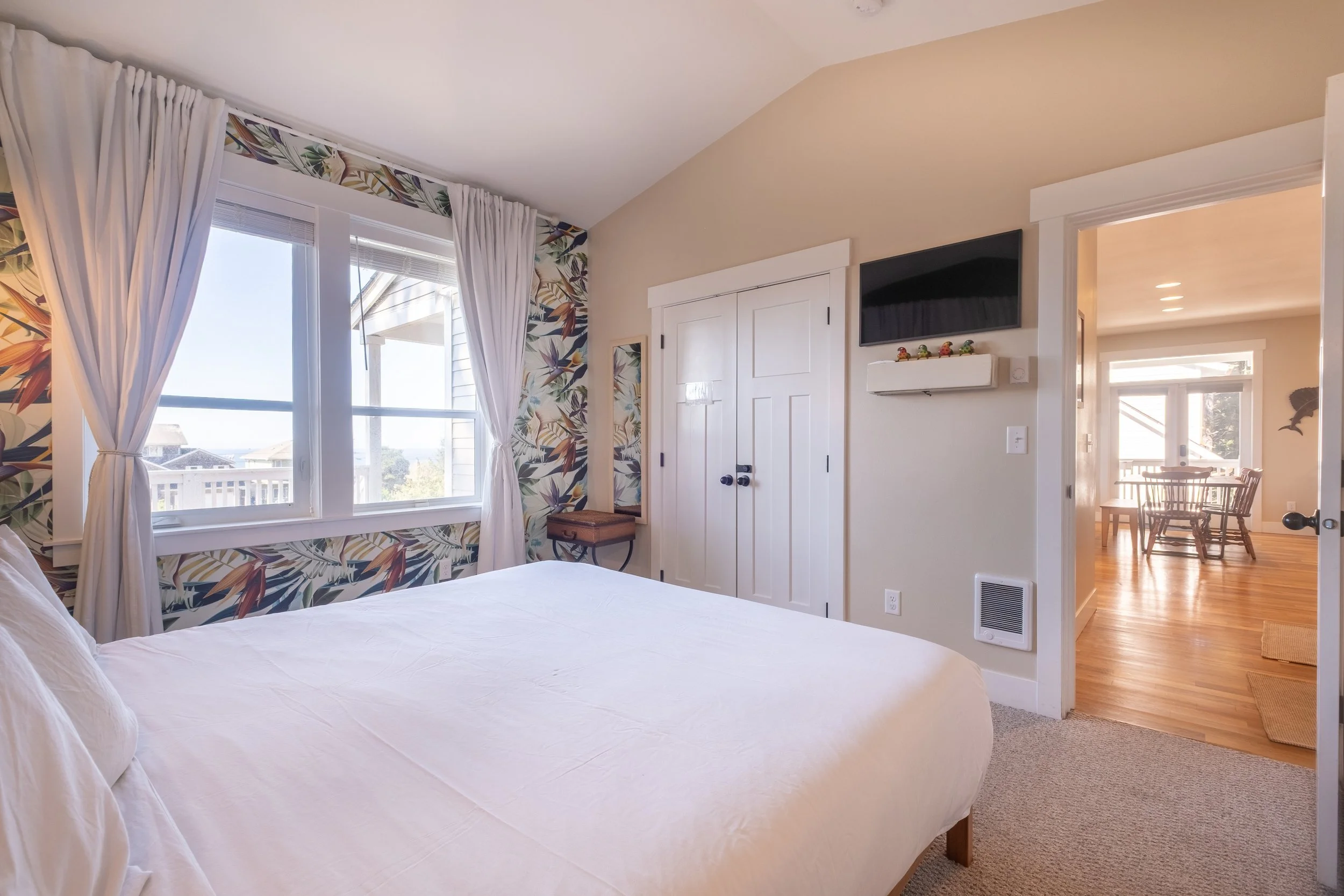 A bedroom with white bedding, a large window with white curtains, floral wallpaper, a small wooden bench, white closet doors, a wall-mounted TV, and a doorway leading to a dining area with a wooden table and chairs.