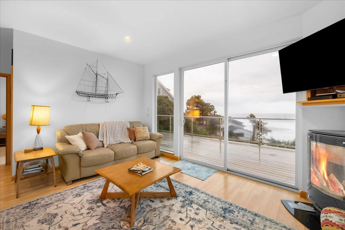 Living room with beige sofa, wooden coffee table, area rug, lamp, wall decor, large glass sliding door leading to balcony with ocean view, fireplace, and wall-mounted TV.