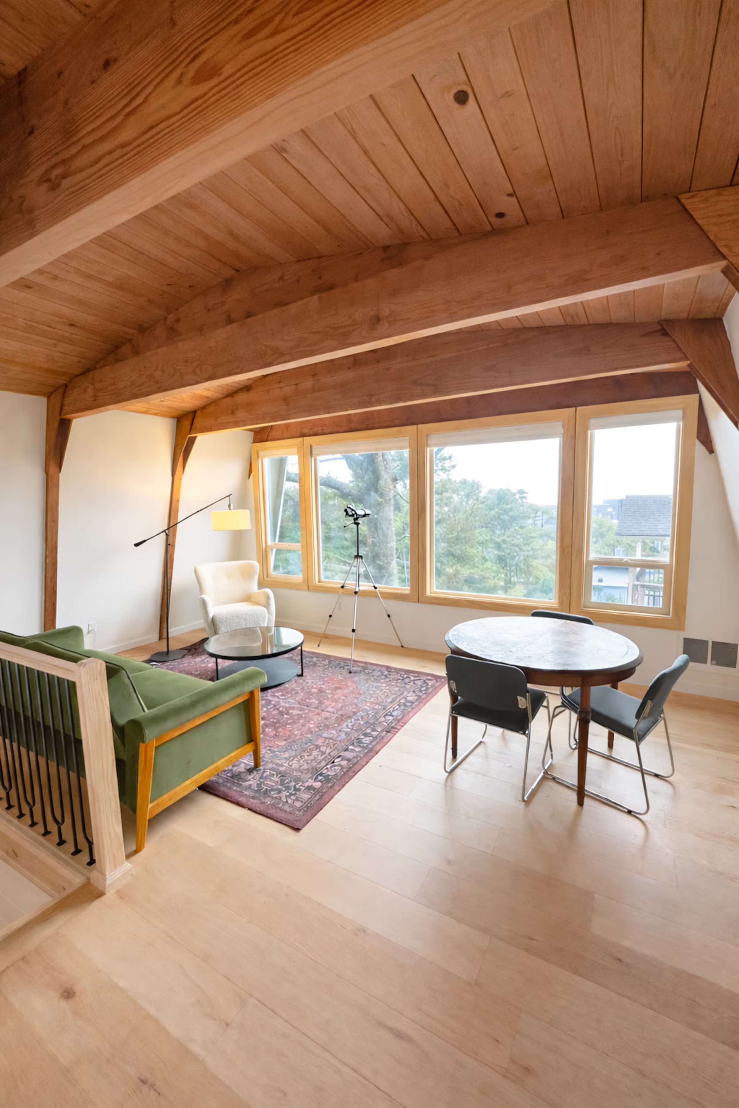 Living room with a wooden ceiling, large windows, a green sofa, a white armchair, a round table, a black chair, a tripod, and a colorful rug on light hardwood flooring.