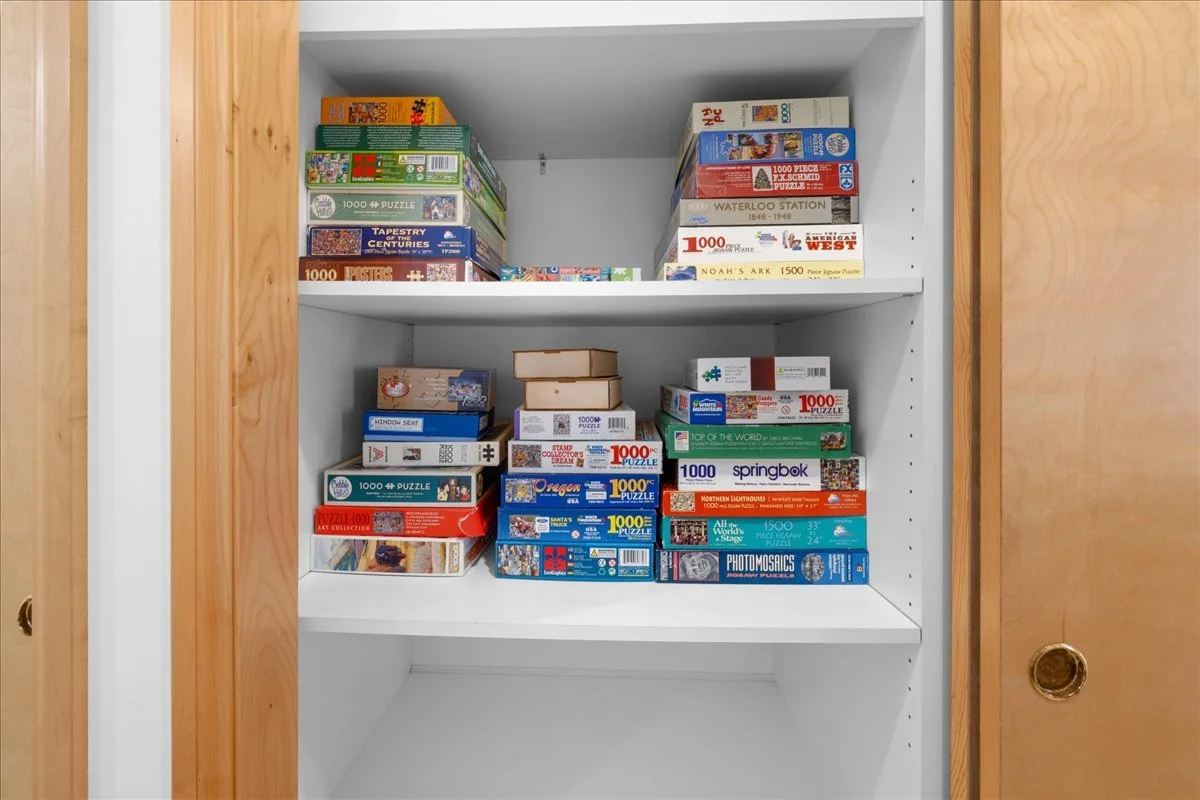 A white bookshelf with three shelves holding multiple puzzle boxes of various sizes and brands, with wooden sliding doors on either side.