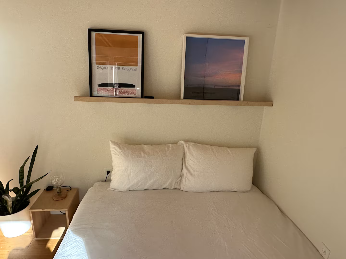 A bedroom with a bed covered in a white textured bedspread, two white pillows, a wooden nightstand with a small lamp and a plant, and two framed pictures on the wall. One picture shows a sunset, and the other has text and a graphic design.