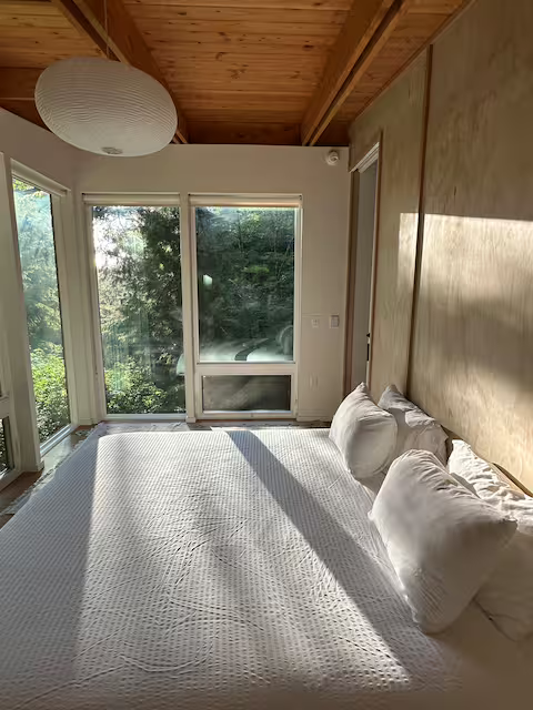 A bedroom with a large bed, white bedding and pillows, wooden ceiling, and a window showing trees outside, bathed in sunlight.
