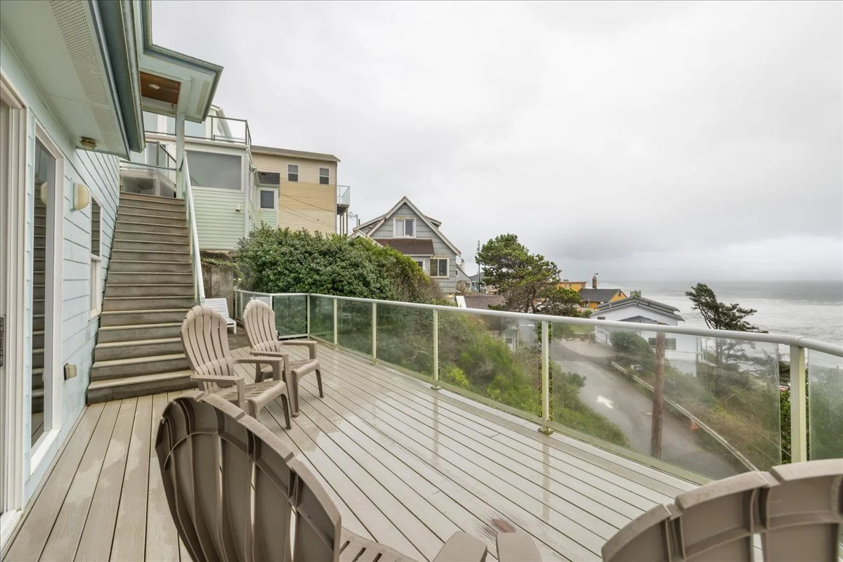 An outdoor wooden deck with seating, overlooking houses, trees, and the ocean on a cloudy day.