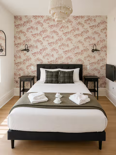 A neatly made bed with white linens, two plaid pillows, and a gray blanket at the foot. Two white towels and decorative items sit on the bed. Two black modern wall lamps are mounted on either side of a pink floral wallpapered wall. A white chandelier